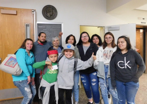 Group photo of smiling White and Native American youth and adults in a school hallway. There is a large circular plaque with a man’s face and a sign behind the group on a white wall. 