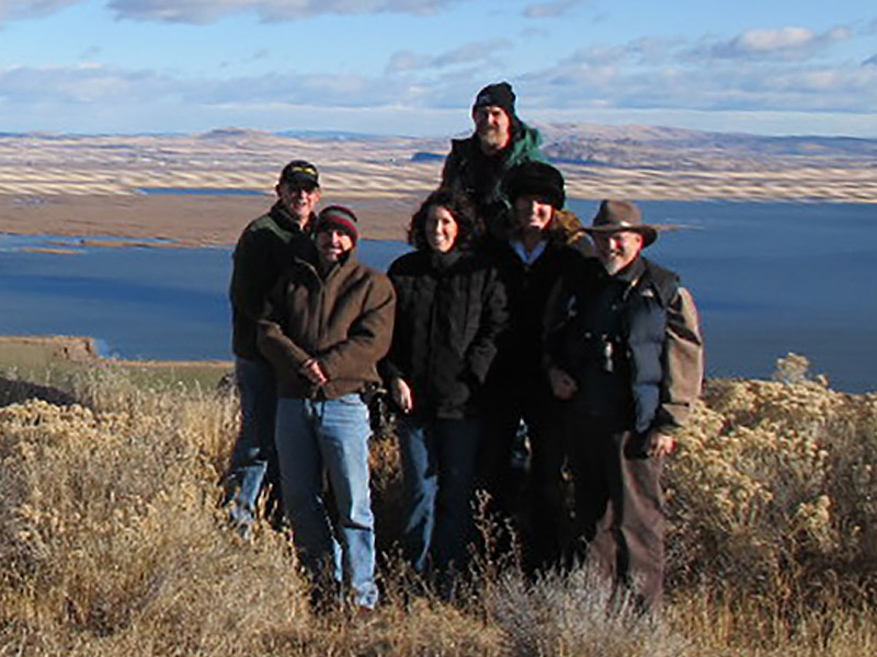Group photo of older men and women smiling with the Klamath River behind them. Everyone is wearing winter jackets and long pants; some are also wearing hats. The group is standing on tall, dry grass. A large body of water and agricultural fields are behind them. 