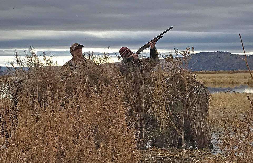 Two men hunting in a valley of tall dry grass. One of the men is pointing a gun toward the sky as if he is about to shoot a bird. 