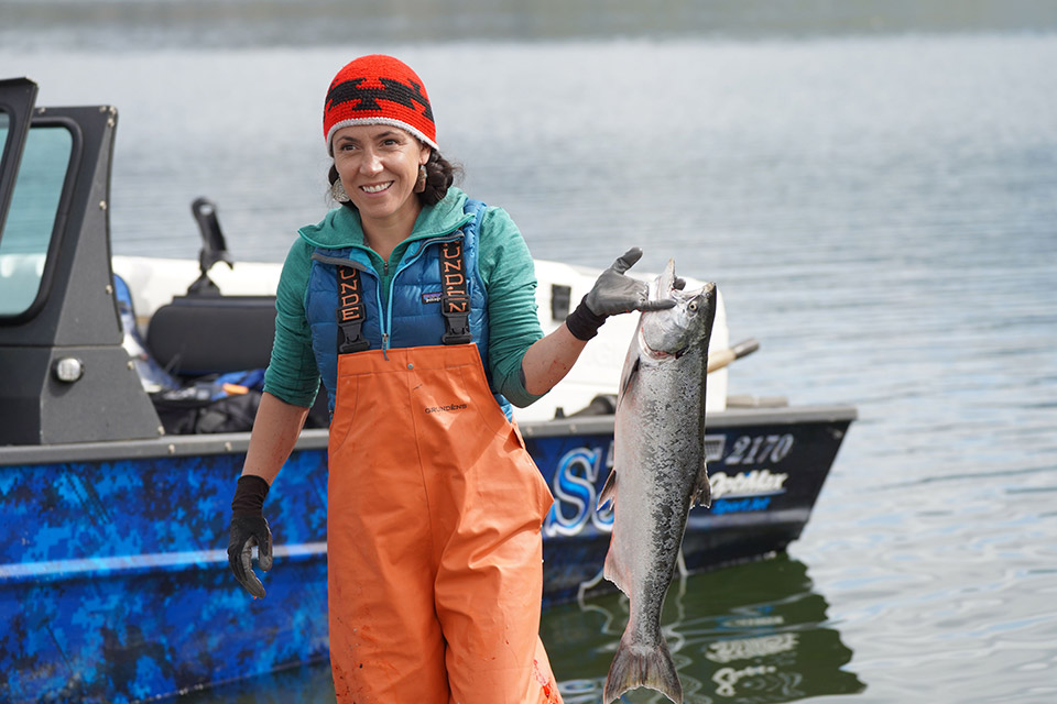 Smiling Native American woman wearing bright orange fishing overalls and a red knit hat holding a salmon. A black and blue boat is on the water behind her.
