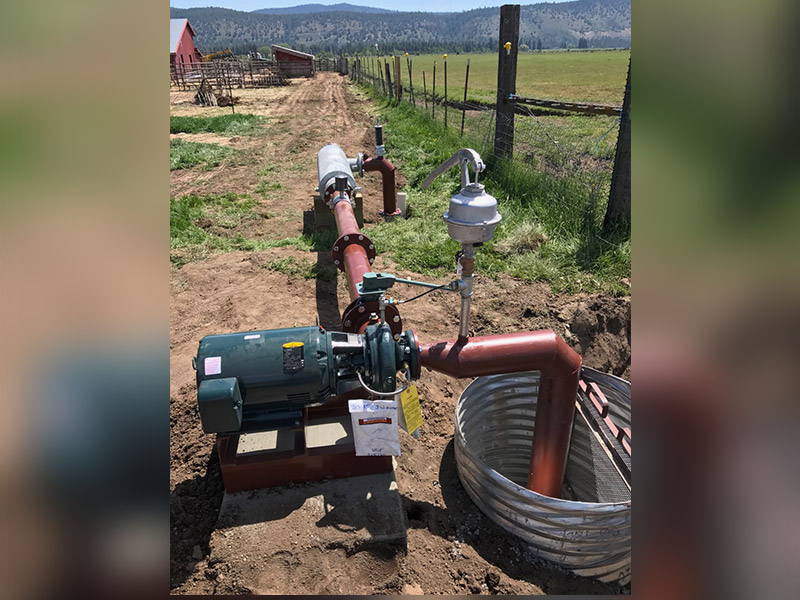 Completed water irrigation system on a farm. The irrigation machinery’s base is dark green, and the pipes are red. Near the irrigation system is an old red wood barn, barbed wire fence and mountains in the background. 