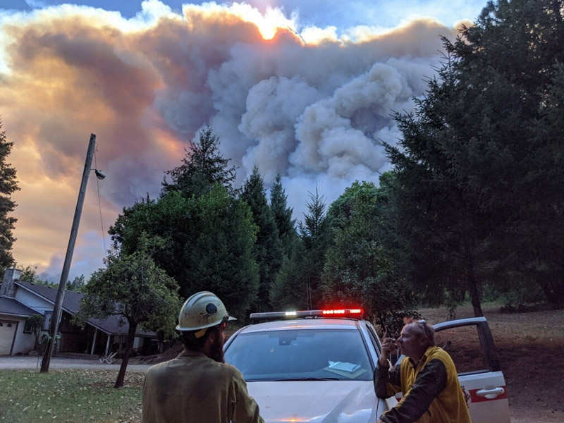 Two emergency responders stand next to a police car with red lights shining on the top of the car. Large plumes of smoke fill the sky behind a home and trees in the background.