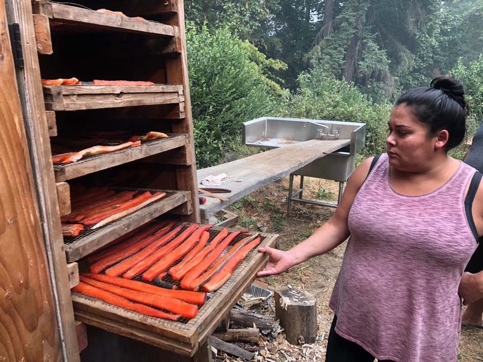 Woman looking at a horizontal rack loaded with strips of salmon. There are 5 racks of salmon visible inside a wooden box used for smoking the fish. The entire scene takes place in an outdoor kitchen in a wooded area.