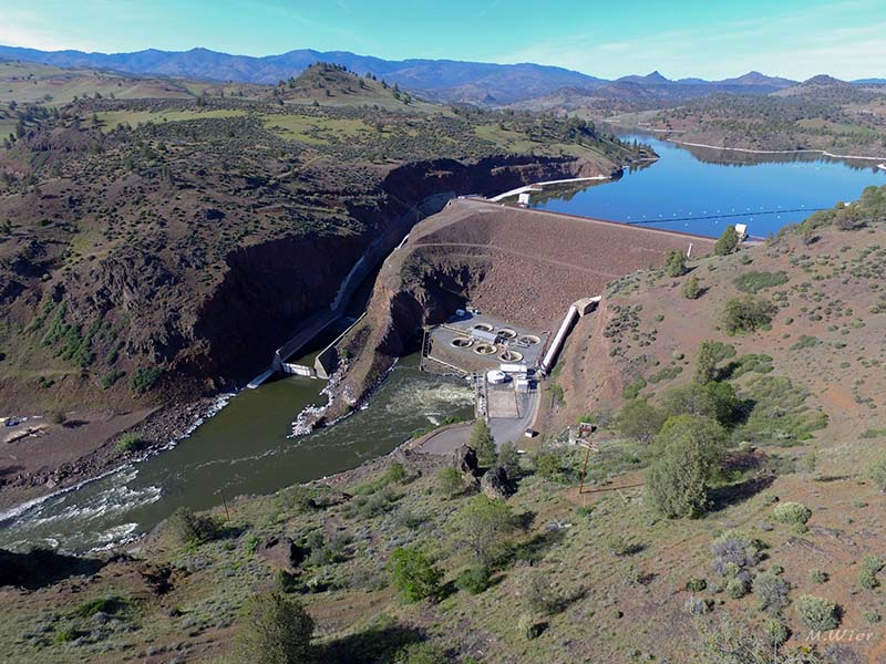 A dam disrupts the flow of a river. Behind the dam is a large reservoir. The hills around the dam are mostly dry and brown but there is some dispersed green trees and shrubs throughout the landscape. A mountain range stands in the background.