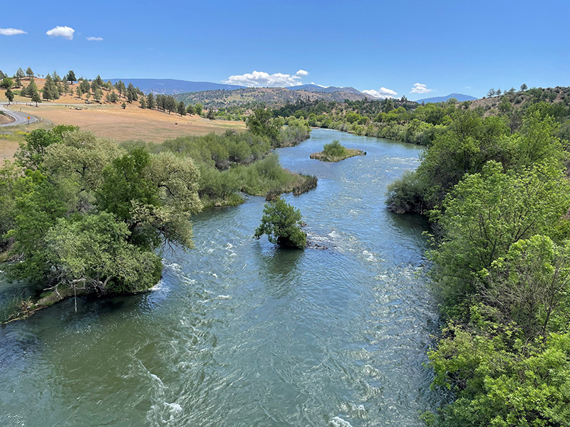 A river twists from the viewer into the distance under a blue sky with scattered clouds on the hilly horizon. Foliage grows along the riverbanks and even on small islets in the middle of the water.